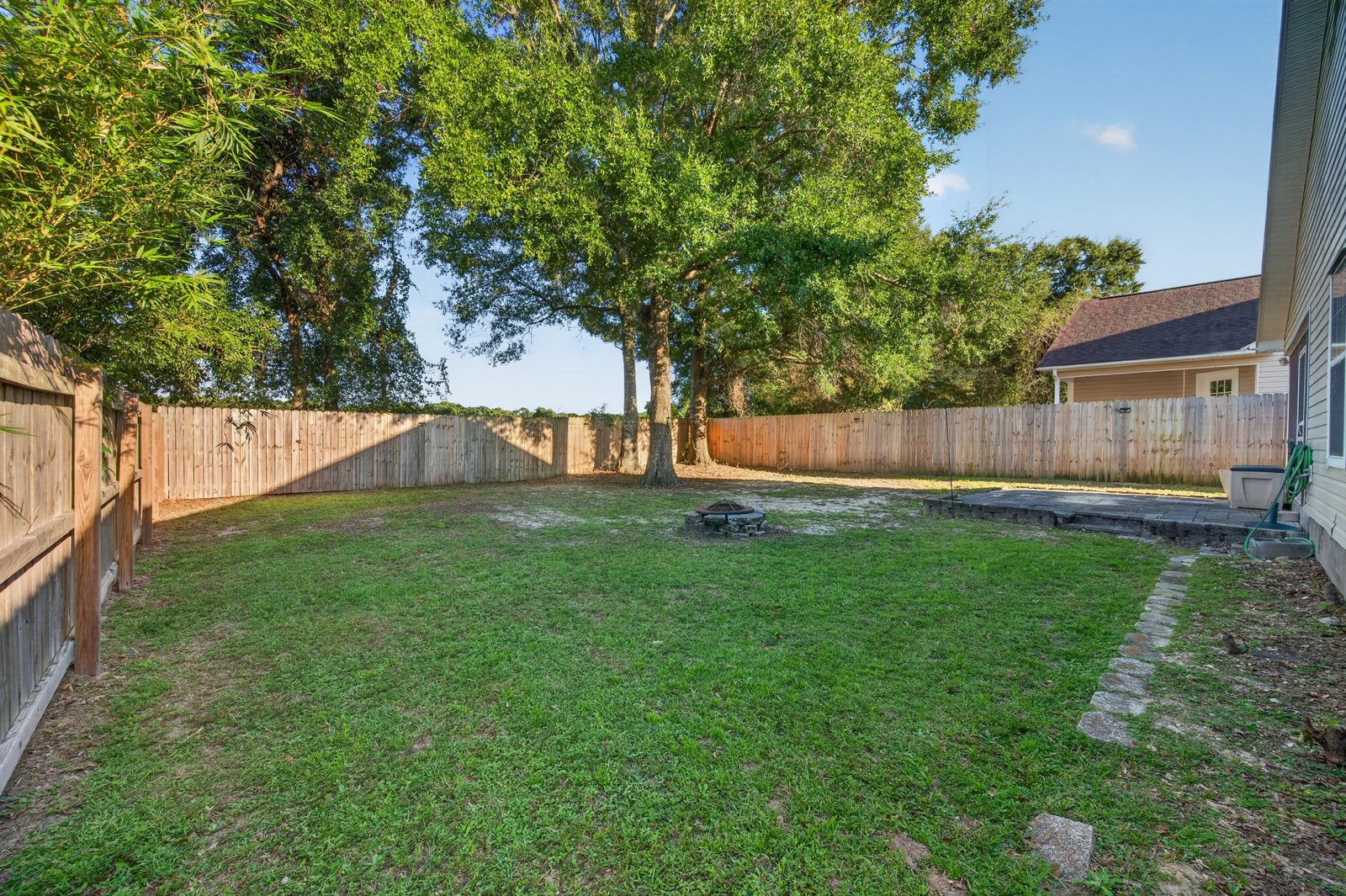Wide view of the privacy-fenced backyard with mature shade trees and a stone path along the house