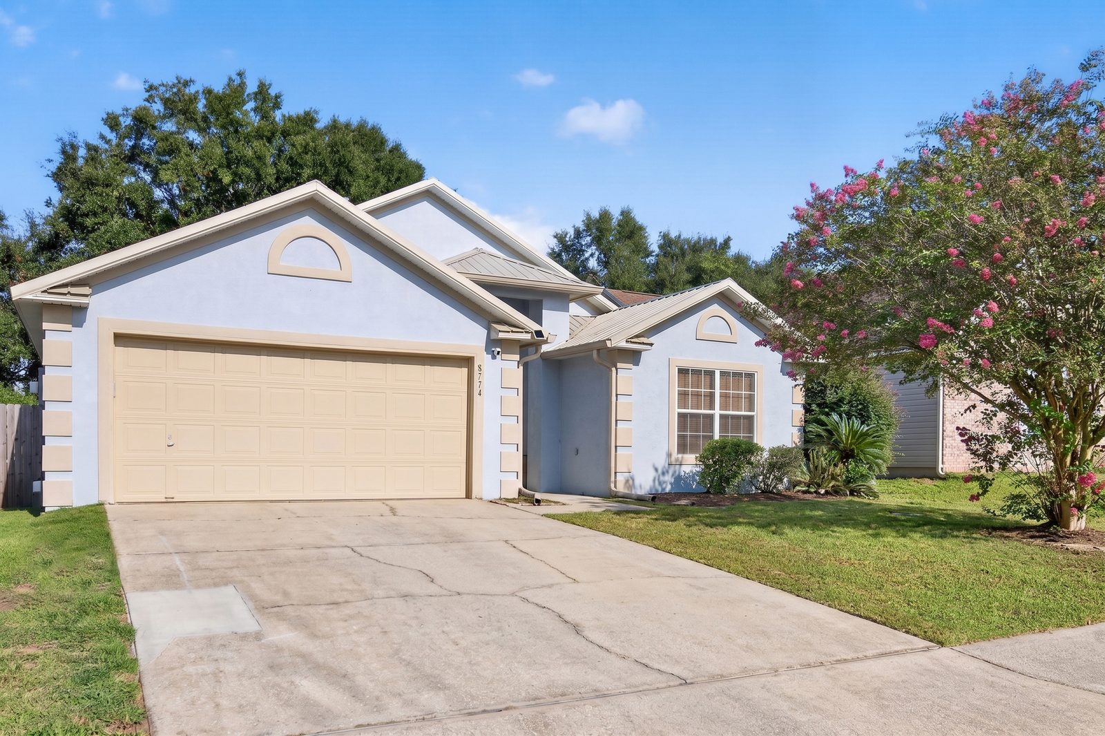 Front of 8224 Lode Star Avenue: a single-story light blue stucco home with a metal roof, a two-car garage, and a flowering crape myrtle by the lawn